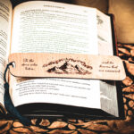 Detailed view of a handcrafted birch wood bookmark with a dark teal tassel, showcasing the mountain peak and celestial engraving across the pages of Iron Flame.