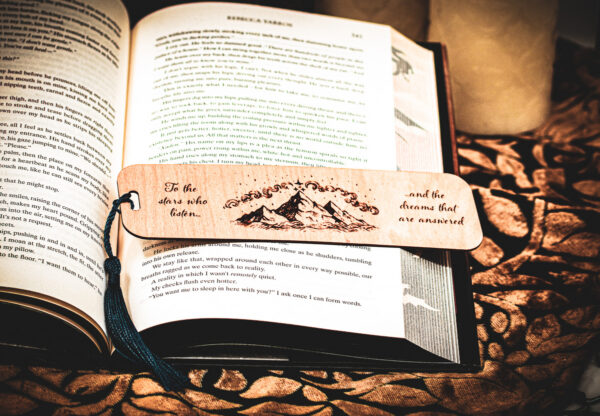 Detailed view of a handcrafted birch wood bookmark with a dark teal tassel, showcasing the mountain peak and celestial engraving across the pages of Iron Flame.