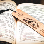 A laser cut and engraved bookmark featuring three mountain peaks with three stars framing the topmost peak with clouds in an arc above them with the words, "To the stars who listen and the dreams that are answered" on either side of the image with a dark blue tassel on top of the page of an open book.
