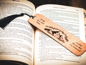 A laser cut and engraved bookmark featuring three mountain peaks with three stars framing the topmost peak with clouds in an arc above them with the words, "To the stars who listen and the dreams that are answered" on either side of the image with a dark blue tassel on top of the page of an open book.