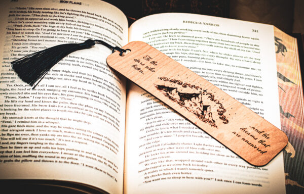 A laser cut and engraved bookmark featuring three mountain peaks with three stars framing the topmost peak with clouds in an arc above them with the words, "To the stars who listen and the dreams that are answered" on either side of the image with a dark blue tassel on top of the page of an open book.
