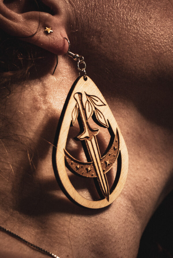 A close-up side profile of a person wearing a Nocturnal Edge earring, showcasing the detailed engraving and natural wood grain against a warm, sunlit background.