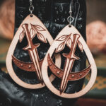 The teardrop dagger earrings styled on a black, ornate palmistry hand model, surrounded by dark academia-themed decor and vintage books.