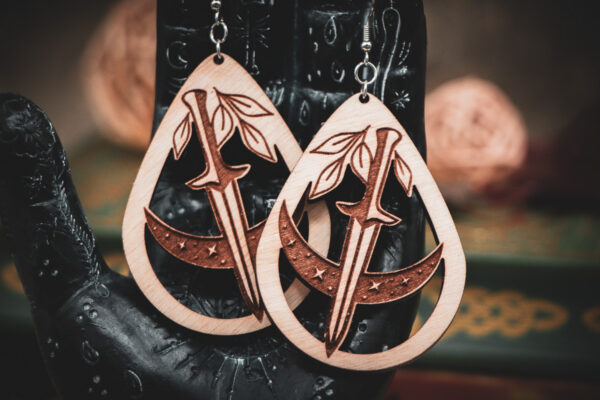 The teardrop dagger earrings styled on a black, ornate palmistry hand model, surrounded by dark academia-themed decor and vintage books.