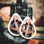 The teardrop dagger earrings styled on a black, ornate palmistry hand model, surrounded by dark academia-themed decor and vintage books.