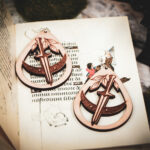 Handcrafted birch wood dagger earrings styled over an antique manuscript featuring whimsical marginalia and drollery illustrations.