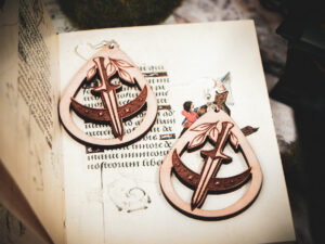 Handcrafted birch wood dagger earrings styled over an antique manuscript featuring whimsical marginalia and drollery illustrations.