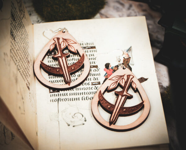 Handcrafted birch wood dagger earrings styled over an antique manuscript featuring whimsical marginalia and drollery illustrations.