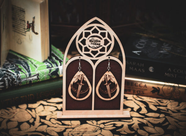 A pair of teardrop-shaped wooden earrings, the "Nocturnal Edge," featuring a laser-cut dagger through a crescent moon with leaf accents, displayed on a custom gothic-arch stand with The Griffin and Stag logo.