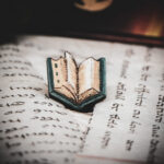 One of the set of three hand painted, laser cut and engraved birch pins in the shape of a Renaissance book in blue sitting on various papers.