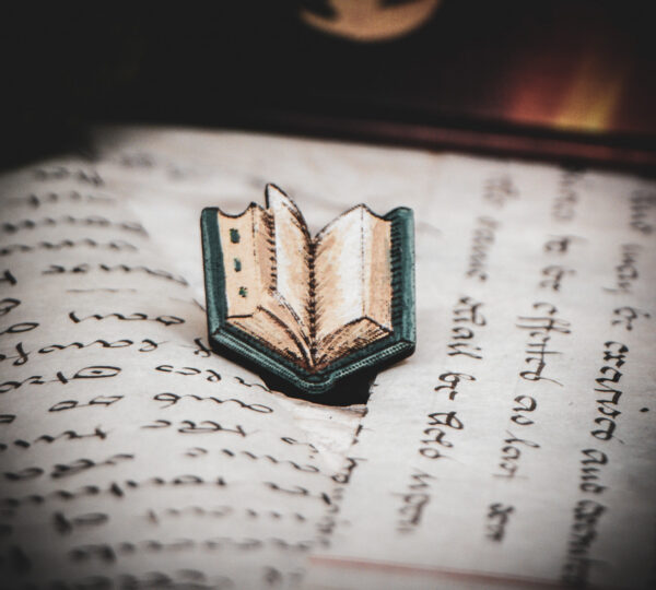 One of the set of three hand painted, laser cut and engraved birch pins in the shape of a Renaissance book in blue sitting on various papers.