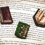 A set of three hand painted, laser cut and engraved birch pins in the shapes of Renaissance books on top of the page of a book.