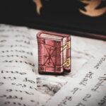 One of the set of three hand painted, laser cut and engraved birch pins in the shape of a Renaissance book in red sitting on various papers.