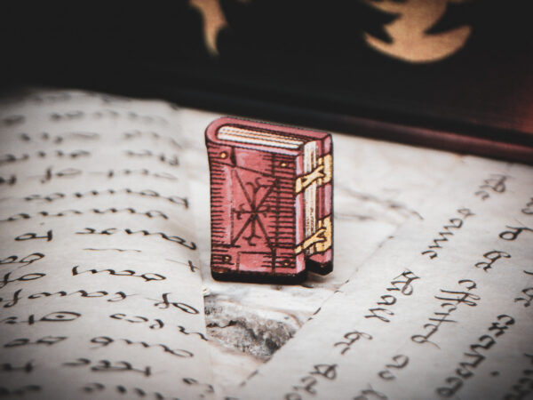 One of the set of three hand painted, laser cut and engraved birch pins in the shape of a Renaissance book in red sitting on various papers.