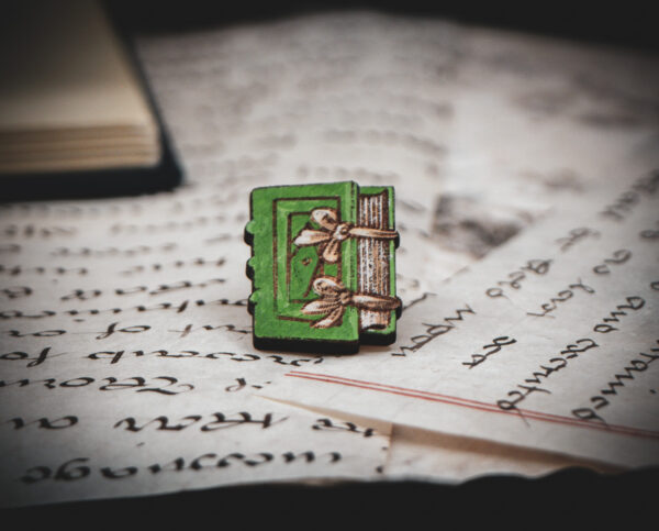 One of the set of three hand painted, laser cut and engraved birch pins in the shape of a Renaissance book in green sitting on various papers.