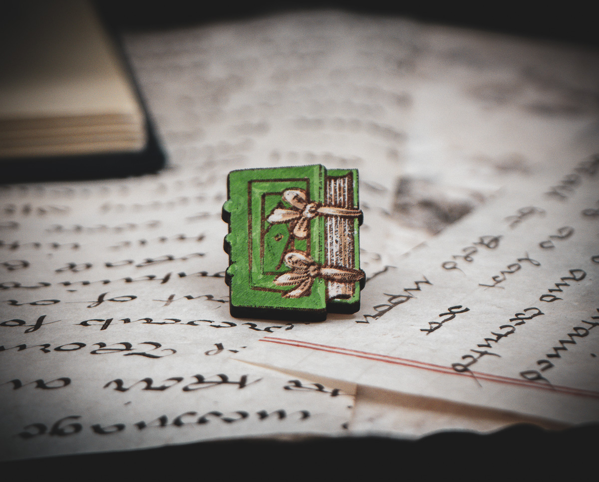 One of the set of three hand painted, laser cut and engraved birch pins in the shape of a Renaissance book in green sitting on various papers.