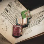 A set of three hand painted, laser cut and engraved birch pins in the shapes of Renaissance books lined up in the middle of two pages of an illuminated manuscript.