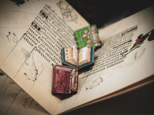 A set of three hand painted, laser cut and engraved birch pins in the shapes of Renaissance books lined up in the middle of two pages of an illuminated manuscript.