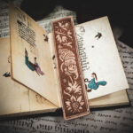 A laser cut and engraved bookmark featuring a creature that looks part-man, part-worm surrounded by leaves and flowers on the page of a small illuminated manuscript.