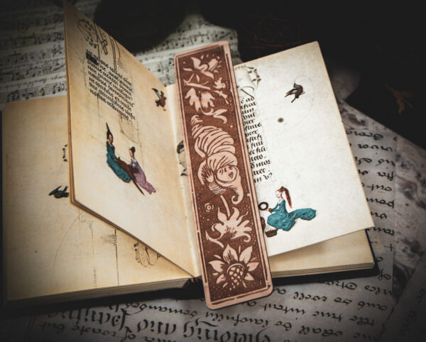 A laser cut and engraved bookmark featuring a creature that looks part-man, part-worm surrounded by leaves and flowers on the page of a small illuminated manuscript.
