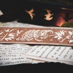 A laser cut and engraved bookmark featuring a creature that looks part-man, part-worm surrounded by leaves and flowers on various papers in front of a stack of books.