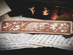 A laser cut and engraved bookmark featuring a creature that looks part-man, part-worm surrounded by leaves and flowers on various papers in front of a stack of books.