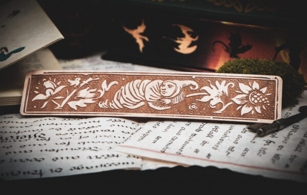 A laser cut and engraved bookmark featuring a creature that looks part-man, part-worm surrounded by leaves and flowers on various papers in front of a stack of books.