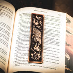 A laser cut and engraved bookmark featuring a creature that looks part-man, part-worm surrounded by leaves and flowers on the page of an open book.