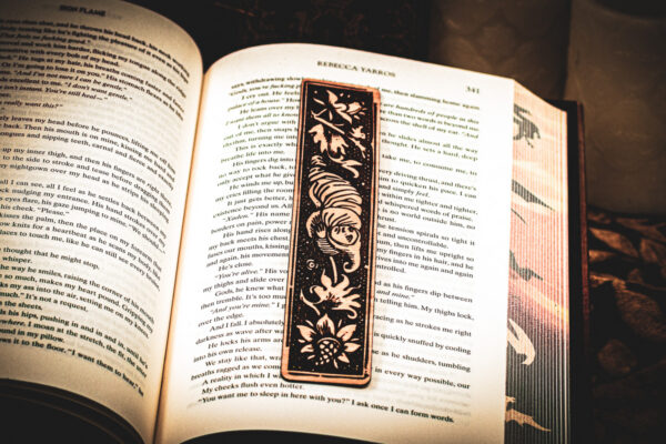 A laser cut and engraved bookmark featuring a creature that looks part-man, part-worm surrounded by leaves and flowers on the page of an open book.