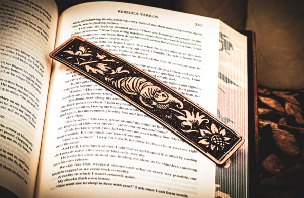 A laser cut and engraved bookmark featuring a creature that looks part-man, part-worm surrounded by leaves and flowers on the page of an open book.