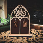 Laser cut and engraved earrings made with 1/16" birch plywood featuring the images of the Tarot cards "Death" and "The World" hanging from a laser cut raw birch stand in the shape of a gothic arch in front of stacks of books.