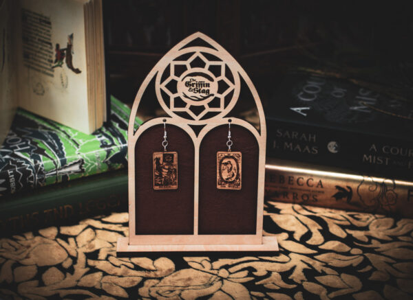 Laser cut and engraved earrings made with 1/16" birch plywood featuring the images of the Tarot cards "Death" and "The World" hanging from a laser cut raw birch stand in the shape of a gothic arch in front of stacks of books.