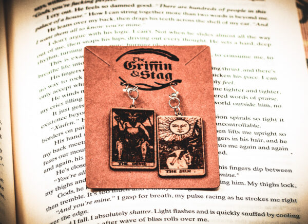 Laser cut and engraved earrings made with 1/16" birch plywood featuring the images of the Tarot cards "The Devil" and "The Sun" hanging from a kraft card on top of the page of a book.