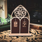 Laser cut and engraved earrings made with 1/16" birch plywood featuring the images of the Tarot cards "The Wheel of Fortune" and "The Tower" hanging from a laser cut raw birch stand in the shape of a gothic arch in front of stacks of books.