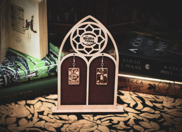Laser cut and engraved earrings made with 1/16" birch plywood featuring the images of the Tarot cards "The Wheel of Fortune" and "The Tower" hanging from a laser cut raw birch stand in the shape of a gothic arch in front of stacks of books.