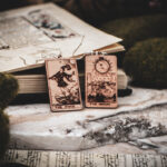 Laser cut and engraved earrings made with 1/16" birch plywood featuring the images of the Tarot cards "The Fool" and "The Moon" leaning against an open illuminated manuscript surrounded by moss rocks on a slab of stone.
