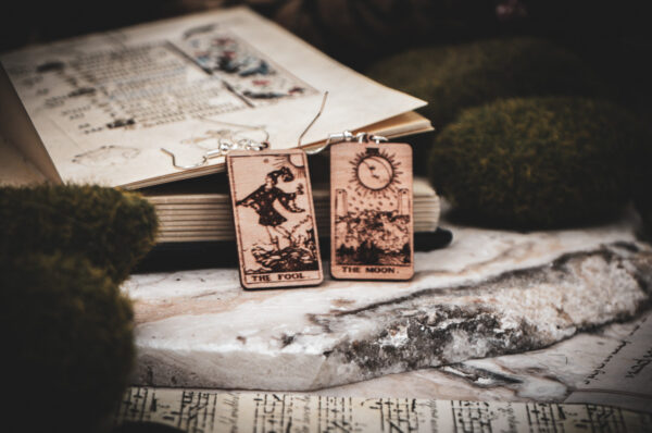 Laser cut and engraved earrings made with 1/16" birch plywood featuring the images of the Tarot cards "The Fool" and "The Moon" leaning against an open illuminated manuscript surrounded by moss rocks on a slab of stone.