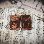 Laser cut and engraved earrings made with 1/16" birch plywood featuring the images of the Tarot cards "The Sun" and "The Devil" on top of papers and a peacock feather on a stone surface.