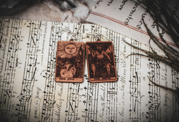Laser cut and engraved earrings made with 1/16" birch plywood featuring the images of the Tarot cards "The Sun" and "The Devil" on top of papers and a peacock feather on a stone surface.