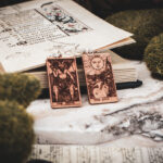Laser cut and engraved earrings made with 1/16" birch plywood featuring the images of the Tarot cards "The Devil" and "The Sun" leaning against an open illuminated manuscript surrounded by moss rocks on a slab of stone.