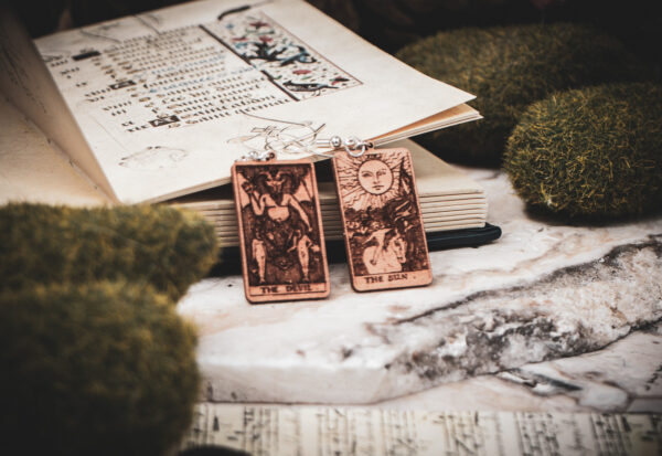 Laser cut and engraved earrings made with 1/16" birch plywood featuring the images of the Tarot cards "The Devil" and "The Sun" leaning against an open illuminated manuscript surrounded by moss rocks on a slab of stone.