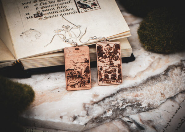 Laser cut and engraved earrings made with 1/16" birch plywood featuring the images of the Tarot cards "The Star" and "Judgement" leaning against an open illuminated manuscript surrounded by moss rocks on a slab of stone.