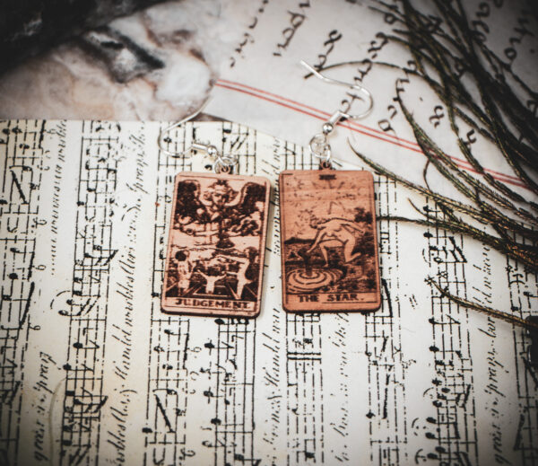 Laser cut and engraved earrings made with 1/16" birch plywood featuring the images of the Tarot cards "Judgement" and "The Star" on top of papers and a peacock feather on a stone surface.
