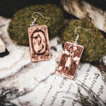 Detailed miniature tarot earrings "The World" and "The Tower" resting against mossy stones near an open antique manuscript atop a marble surface styled with pages of calligraphy and a peacock feather.