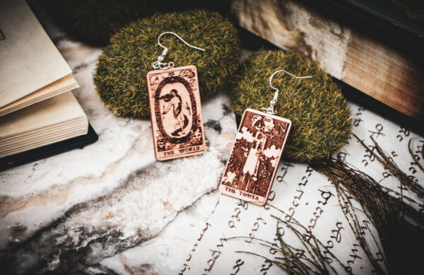 Detailed miniature tarot earrings "The World" and "The Tower" resting against mossy stones near an open antique manuscript atop a marble surface styled with pages of calligraphy and a peacock feather.