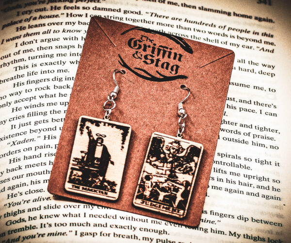 Laser cut and engraved earrings made with 1/16" birch plywood featuring the images of the Tarot cards "The Magician" and "Judgement" hanging from a kraft card on top of the page of a book.