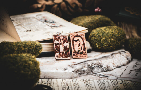 Detailed miniature tarot earrings such as "The Tower" and "The World" resting against an open antique manuscript with whimsical drollery illustrations surrounded by mossy stones on a marble surface.