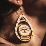A close-up profile of a person wearing a "Seer's Scryer" earring, featuring a laser-engraved crystal ball with a central "all-seeing eye" design on birch plywood.