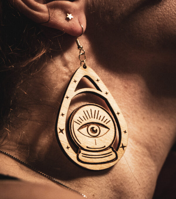 A close-up profile of a person wearing a "Seer's Scryer" earring, featuring a laser-engraved crystal ball with a central "all-seeing eye" design on birch plywood.