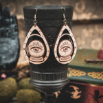 A pair of wooden teardrop-shaped earrings depicting a crystal ball and eye, hanging from a dark metal chalice in a dark academia setting.